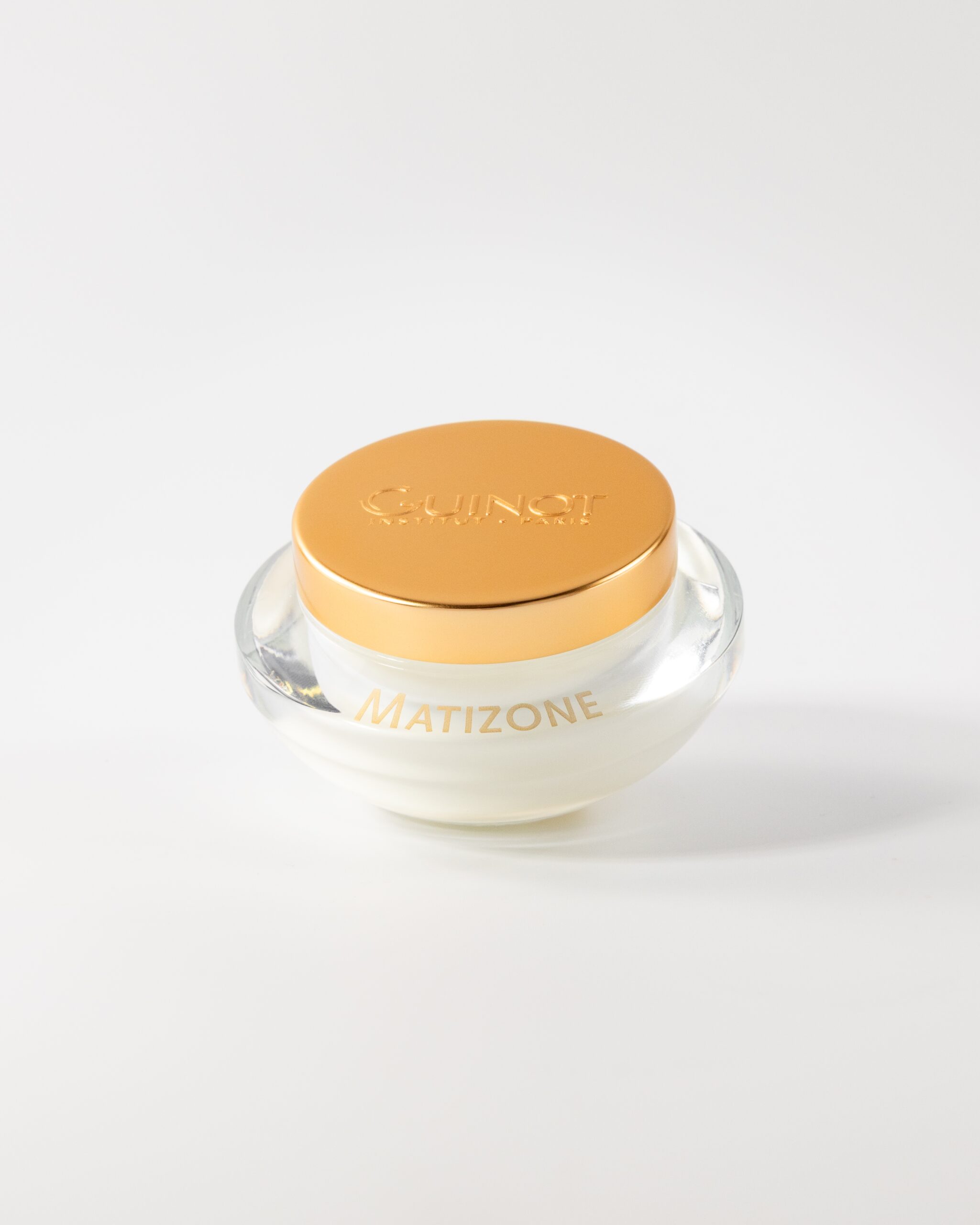 Close-up of a clear cosmetic jar with a metallic orange lid reading 'Guinot Institut Paris' and 'Matizone' on the glass, against a white background.