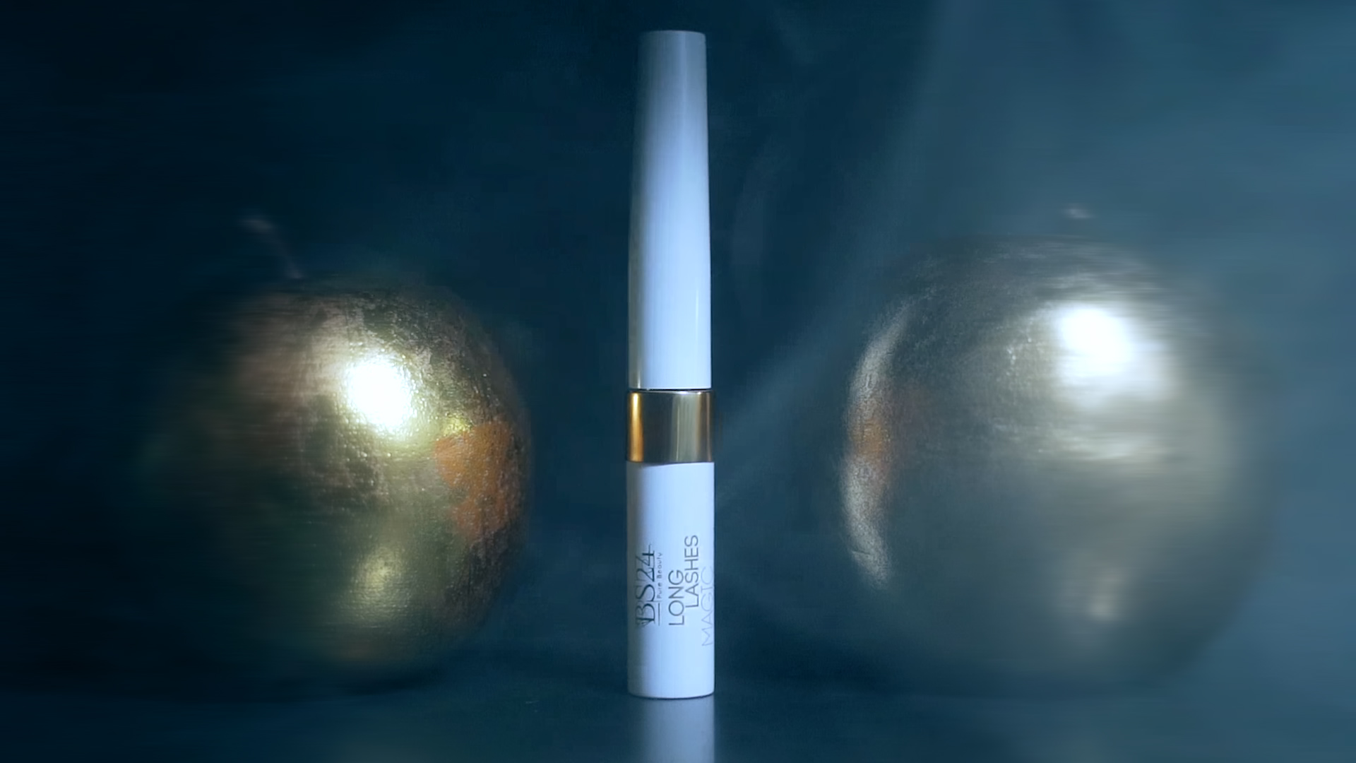 White mascara tube with a gold band stands upright on a dark surface against a moody blue blurred background.
