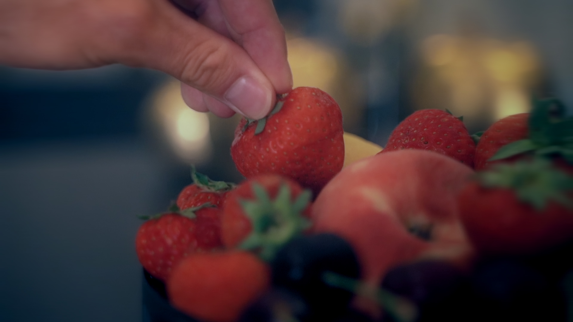Close-up of a hand picking a ripe strawberry from a bowl of mixed berries on a dark surface, focus on the fruit.