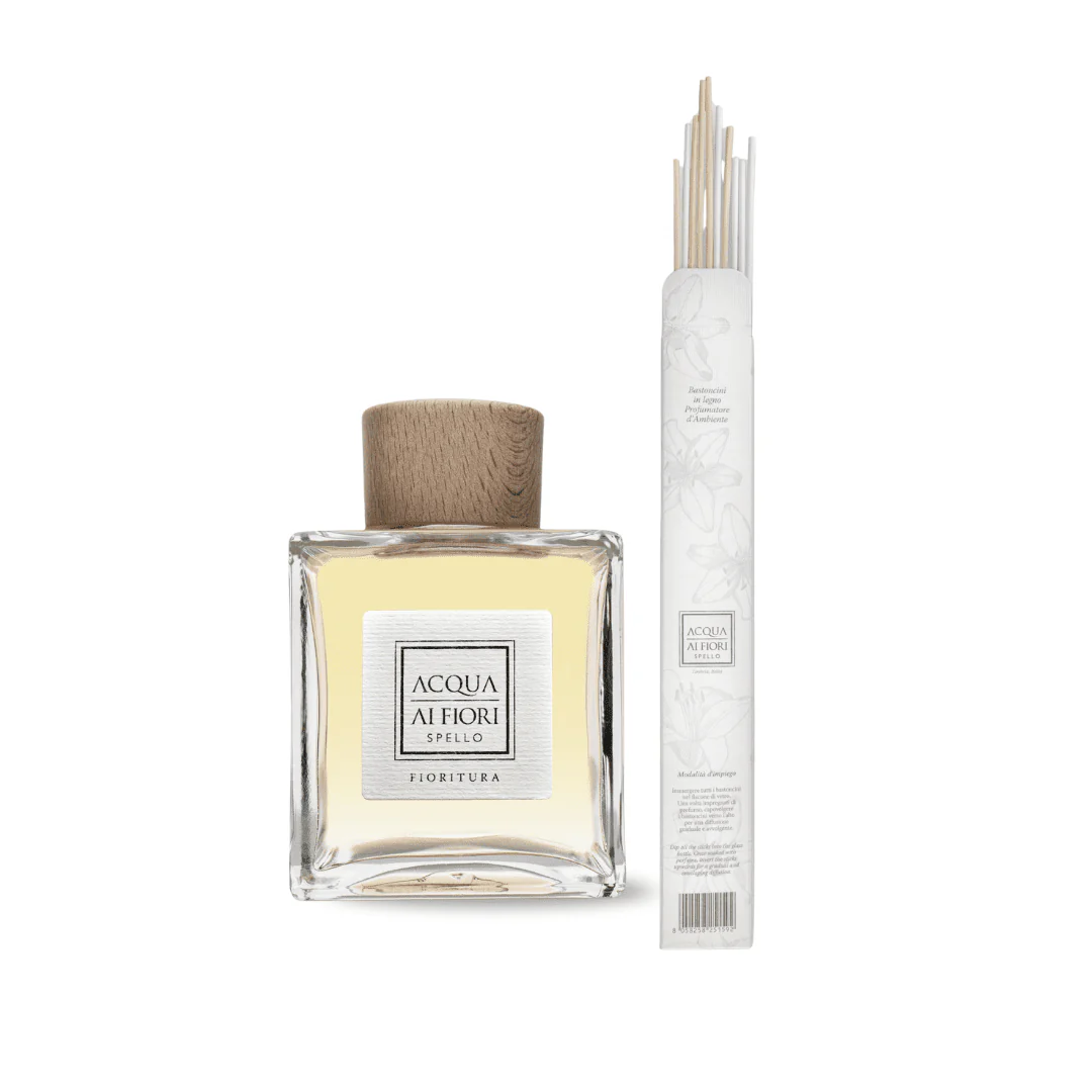 Clear glass perfume bottle with a light yellow liquid and a wooden cap, beside a bundle of white diffuser sticks in a tall white package.