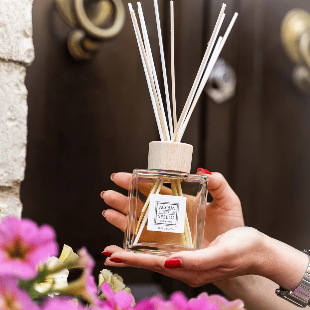 Hand holding a square glass reed diffuser with white sticks and a wooden cap; label reads Acqua di Fiori di Spello.