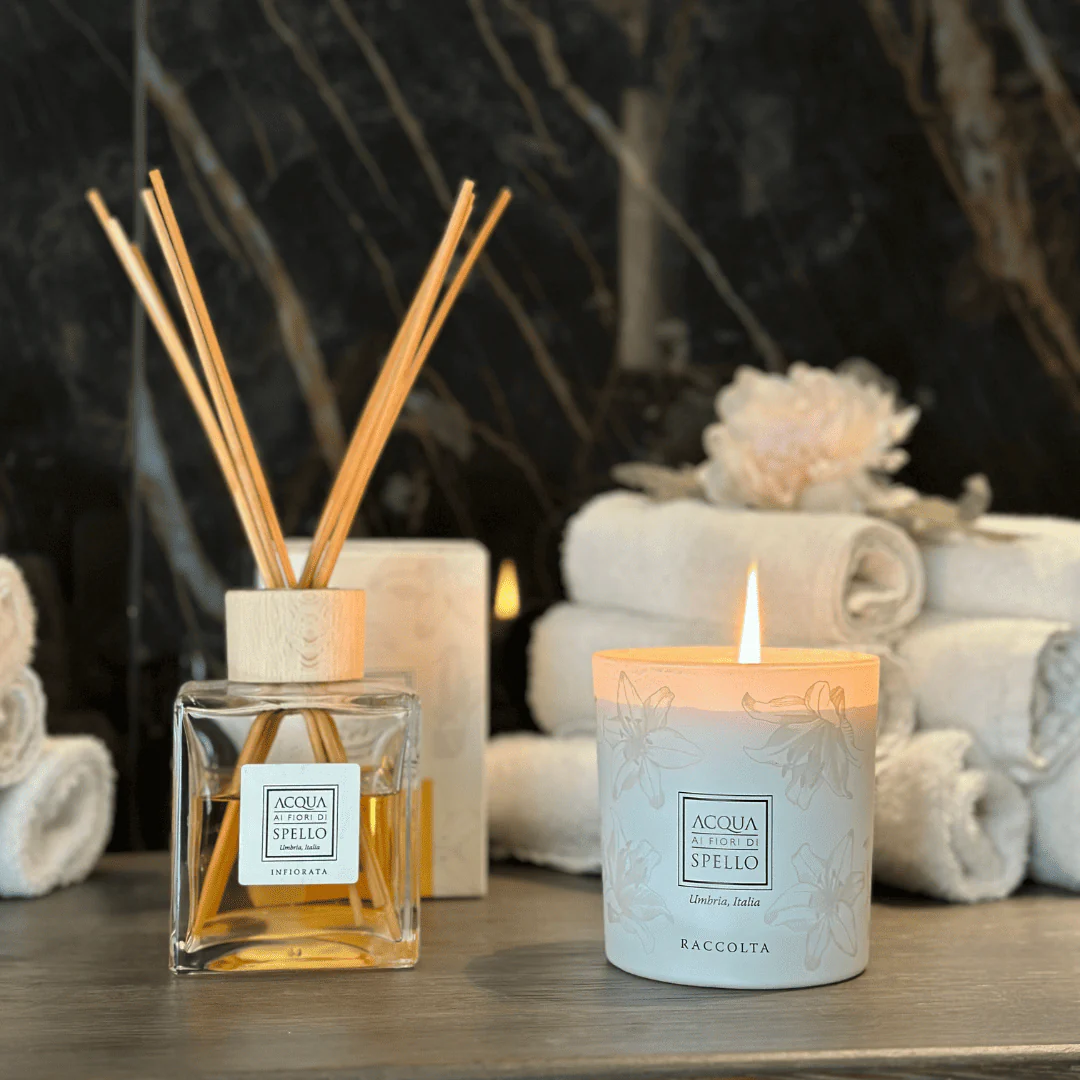 A lit scented candle and a reed diffuser on a wooden surface in a spa setting, with rolled towels in the background.
