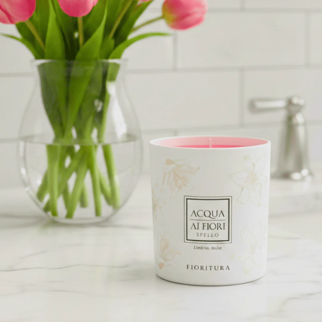 White scented candle with pink wax in a white floral-patterned container on a marble countertop, pink tulips in a glass vase in the background.
