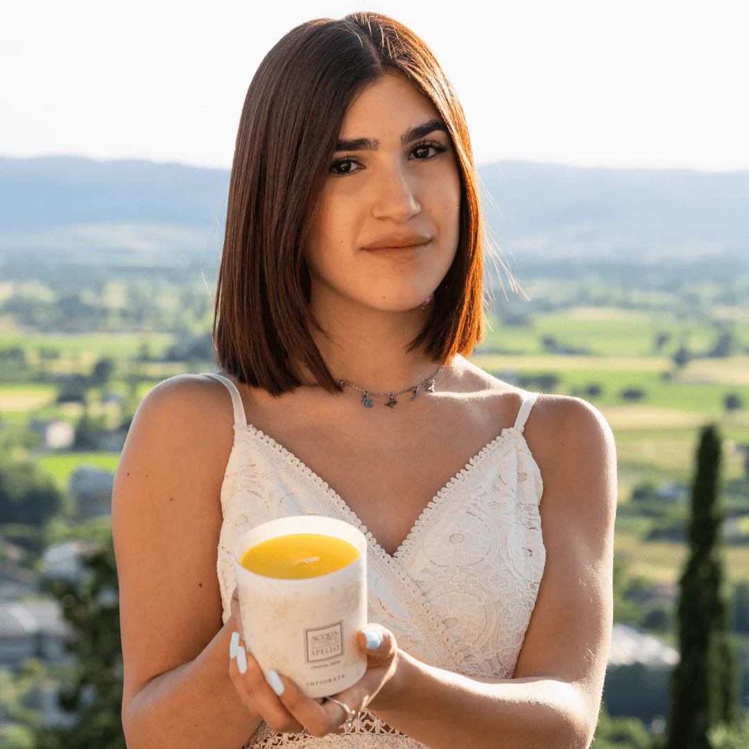 Portrait of a woman with shoulder-length brown hair in a white lace top, holding a lit candle in a decorative mug outdoors.
