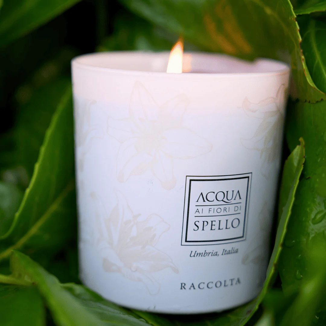 White scented candle with a floral pattern and a lit wick, labeled Acqua ai Fiori di Spello, Umbria, Italy (Raccolta).
