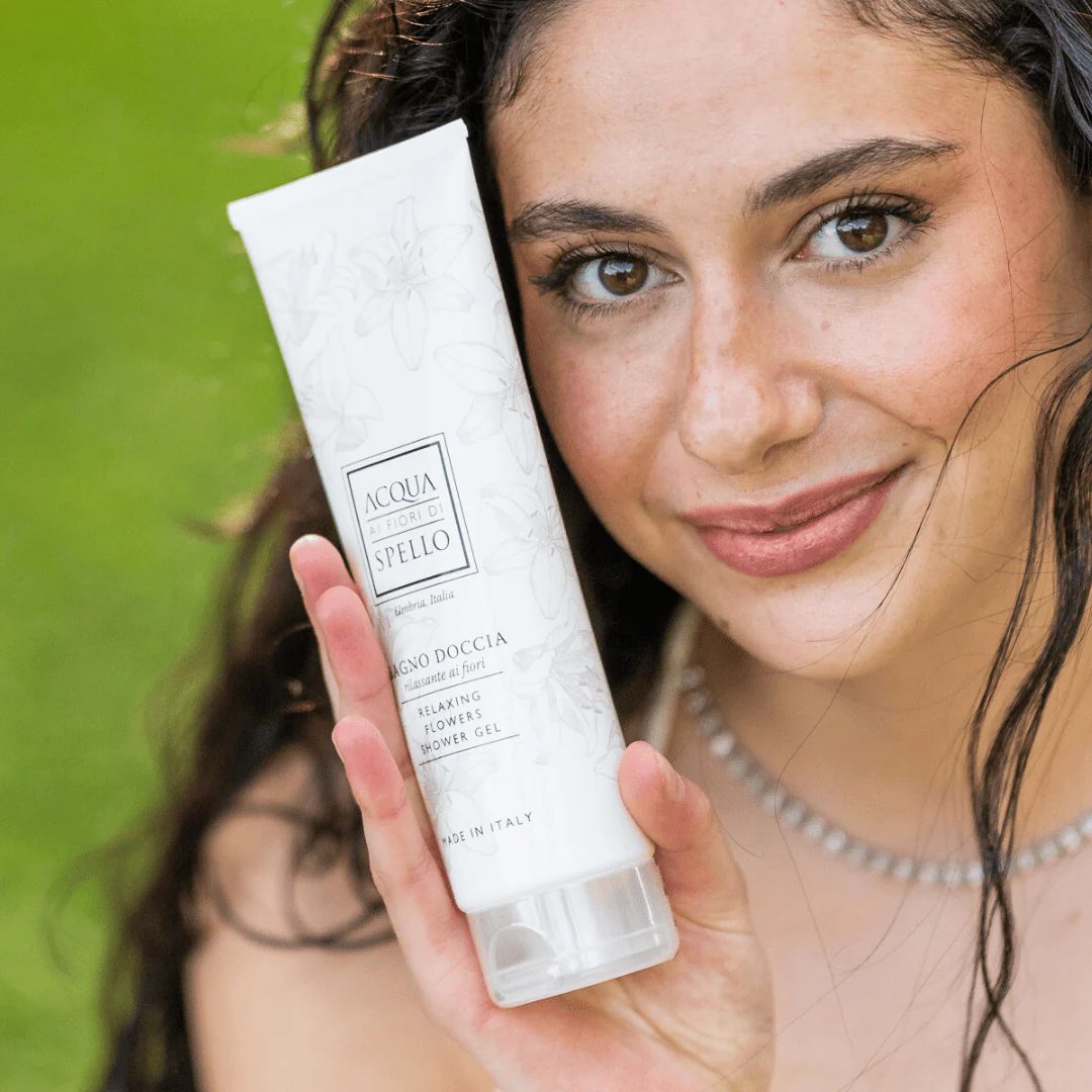 Smiling woman outdoors holds a white floral-patterned tube of shower gel toward the camera.