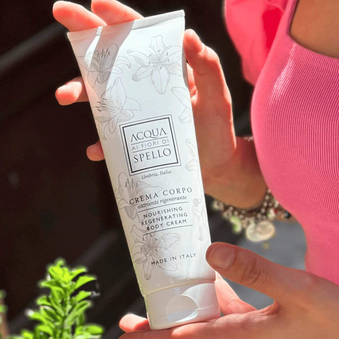 Tube of Nourishing Regenerating Body Cream labeled 'Acqua ai Fiori di Spello' being held by a person in a pink top, Made in Italy