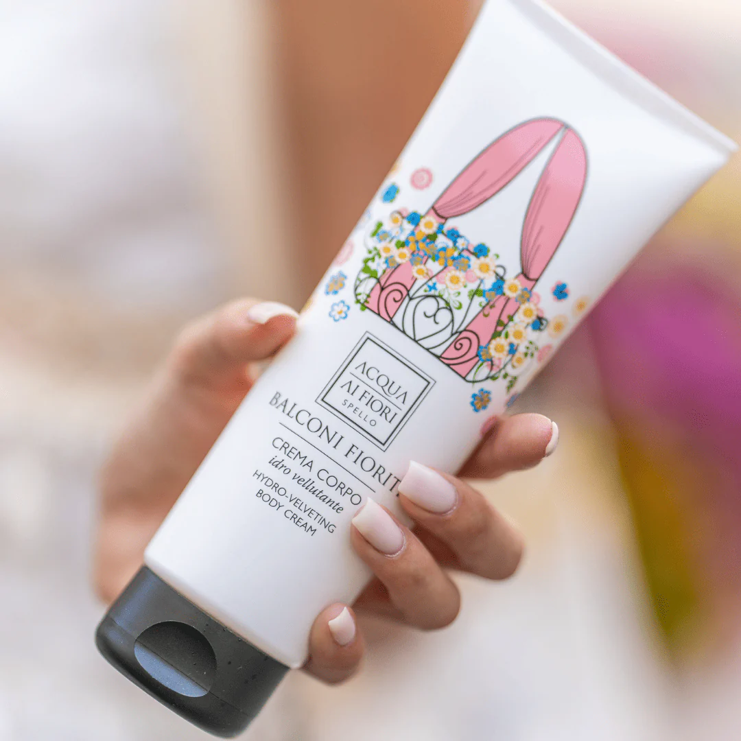 Hand holding a white tube of body cream with colorful floral artwork and a black cap, held near the camera lens.