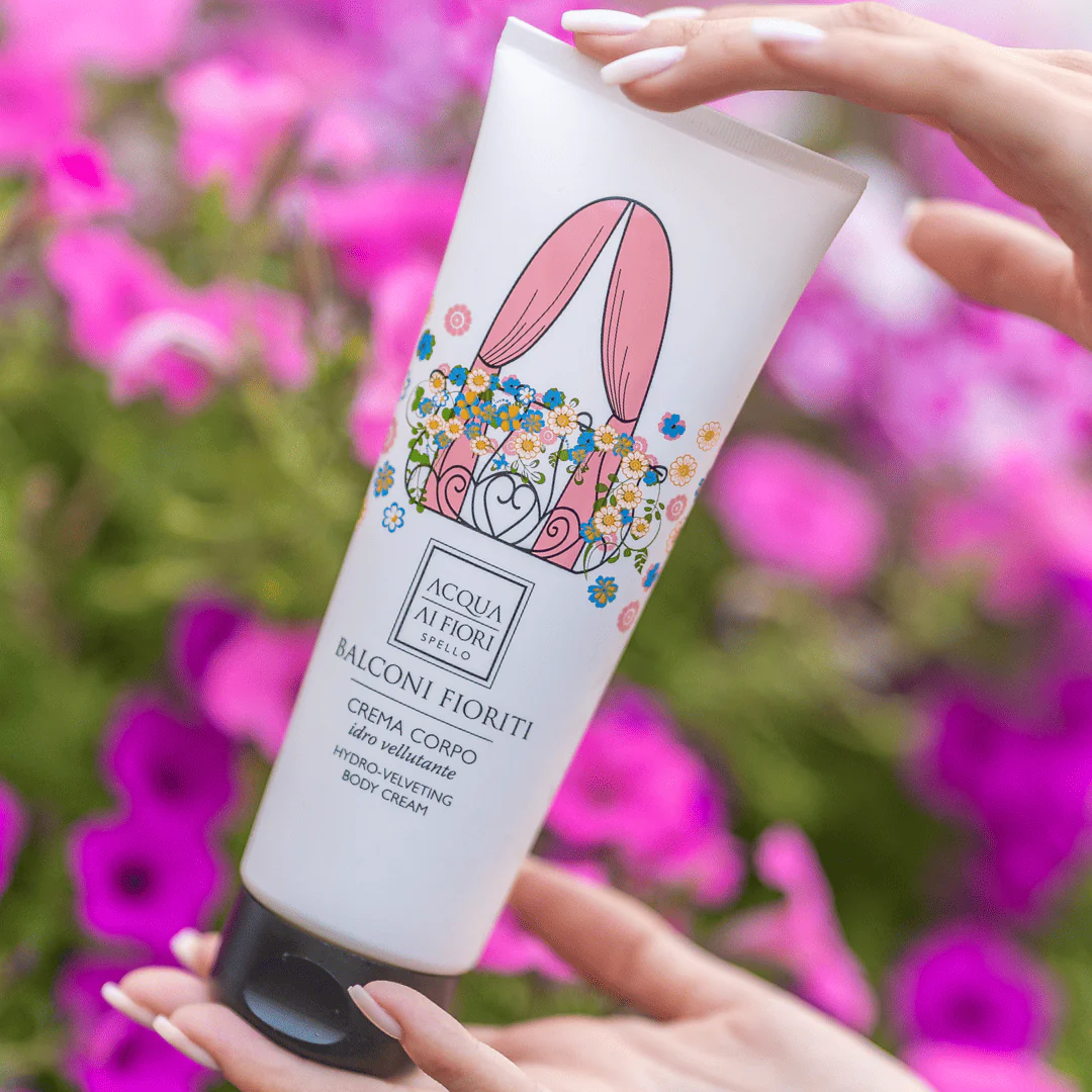 Tube of Balconi Fioriti hydro-velvet body cream being held by hands with a colorful floral design, pink garden flowers in the background