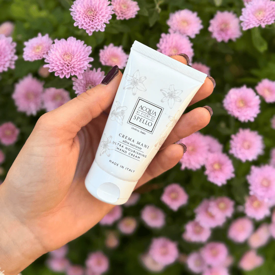 Hand holding a white tube of Crema Mani hand cream with floral illustrations and pink blossoms in the background.
