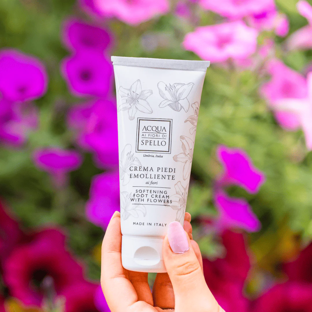 Tube of Acqua ai Fiori di Spello softening foot cream held in front of blurred pink flowers in the background (hand visible).