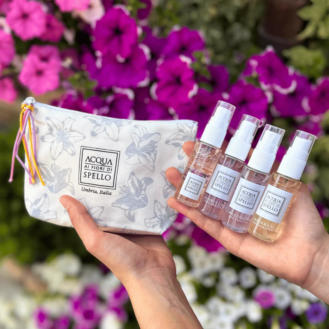 Hands hold a white floral zipper pouch and five Acqua di Spello perfume spray bottles against pink flowers.