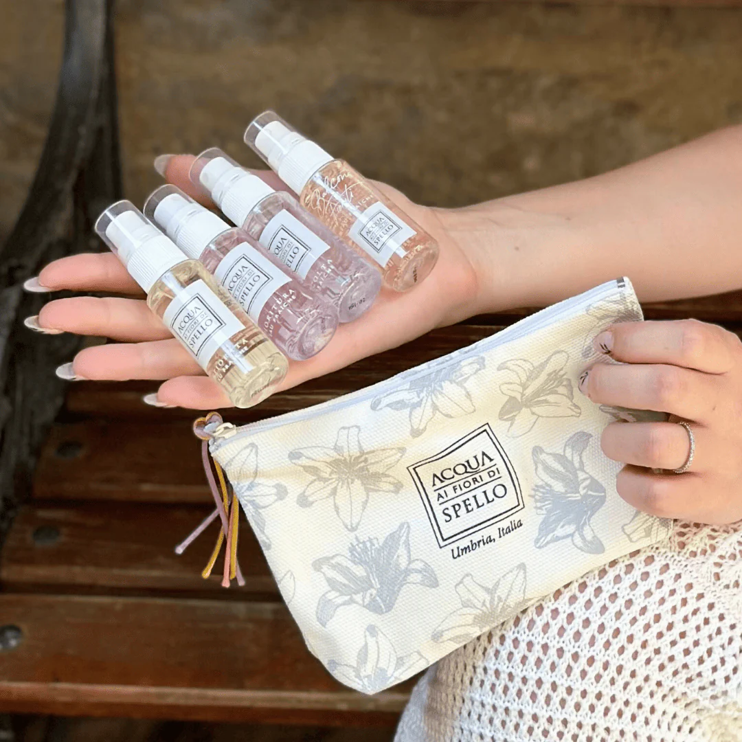 Five small spray bottles with clear liquids held in a hand above a floral-patterned pouch.