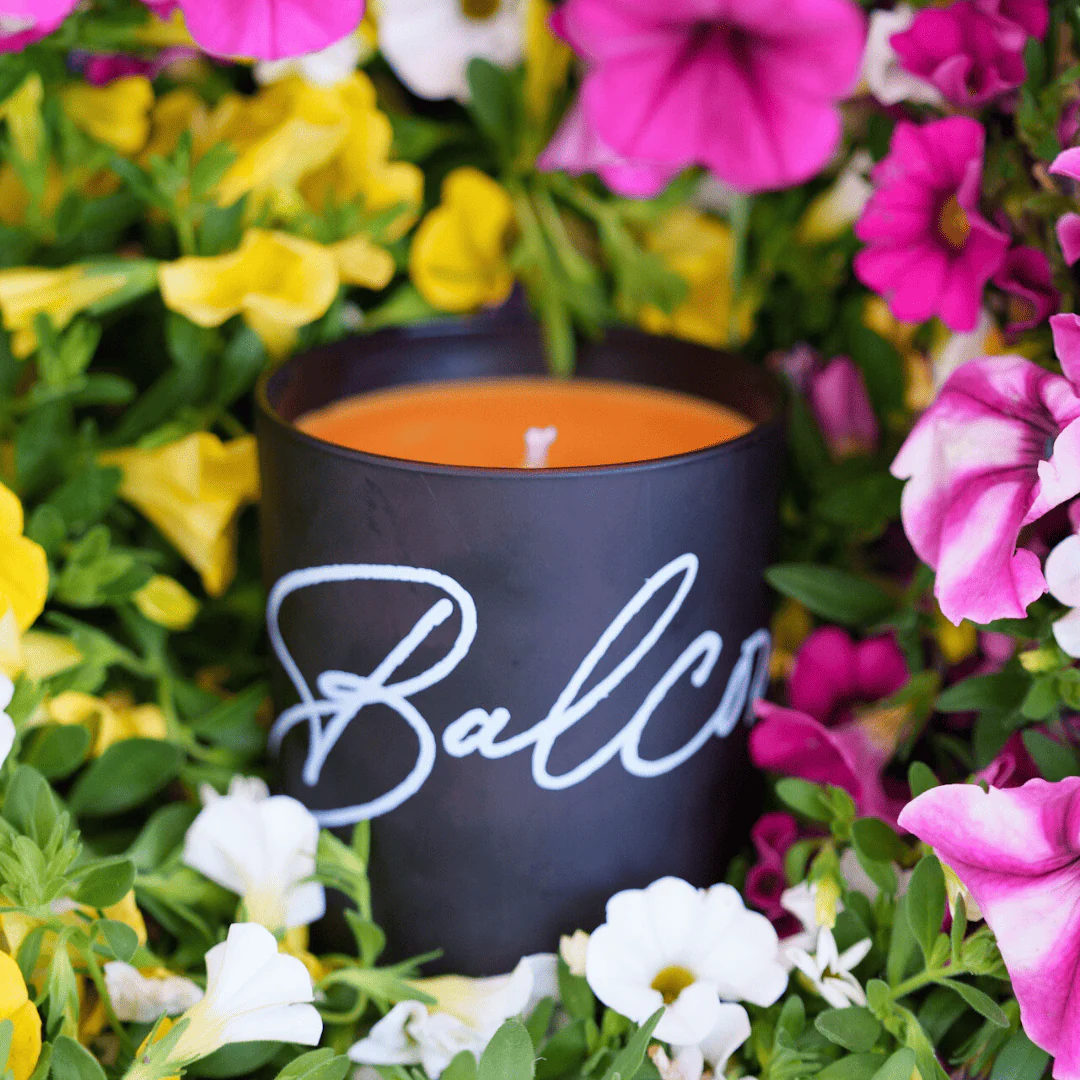 Orange candle in a black container with white cursive writing, nestled among bright pink, yellow, and white flowers.