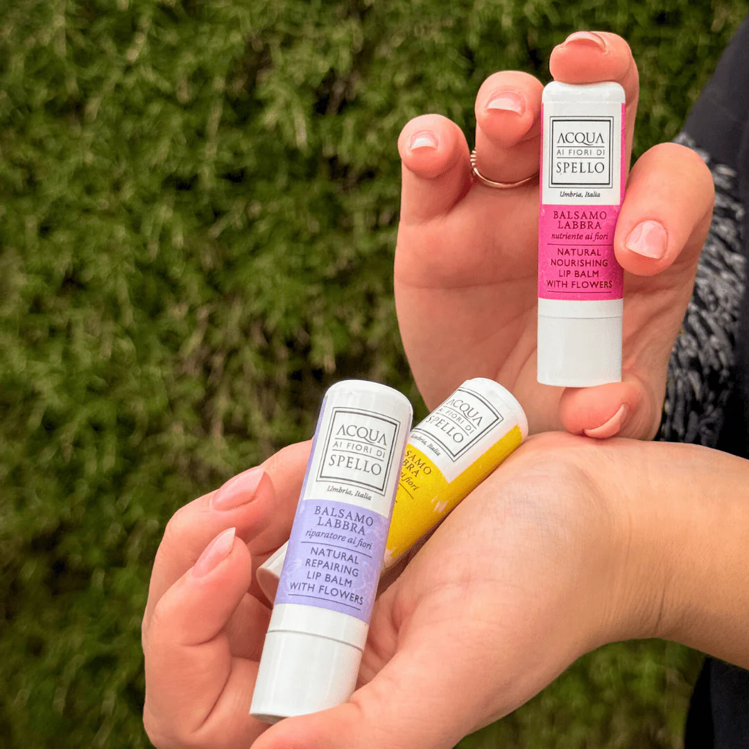 Three Acqua di Spello lip balms in pink, yellow and purple labels held by hands outdoors