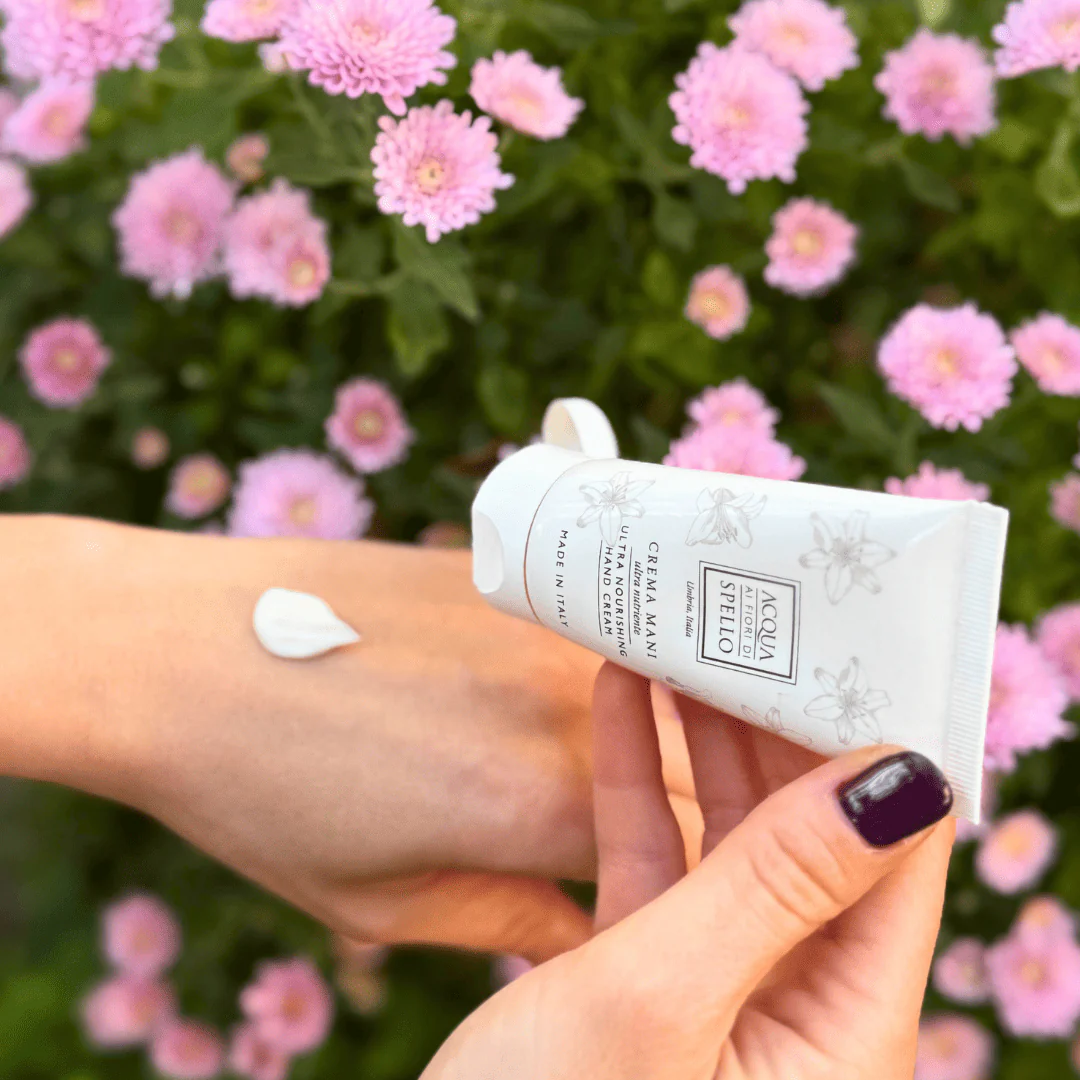 Close-up of a hand applying lotion from a white tube onto the inner forearm, pink flowers in the background.